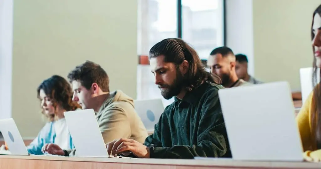 A Caucasian male student asking a question to the professor. Other students are seated