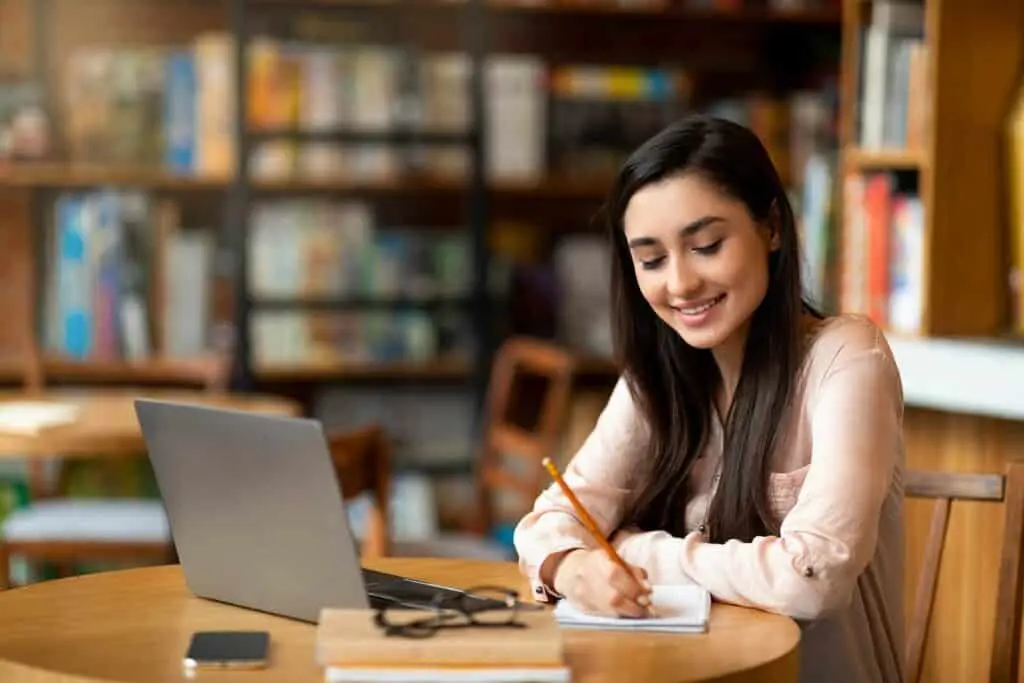Online education. Motivated latin female studying with laptop at cafe and taking notes while watching webinar or preparing for exam in cafeteria, empty space