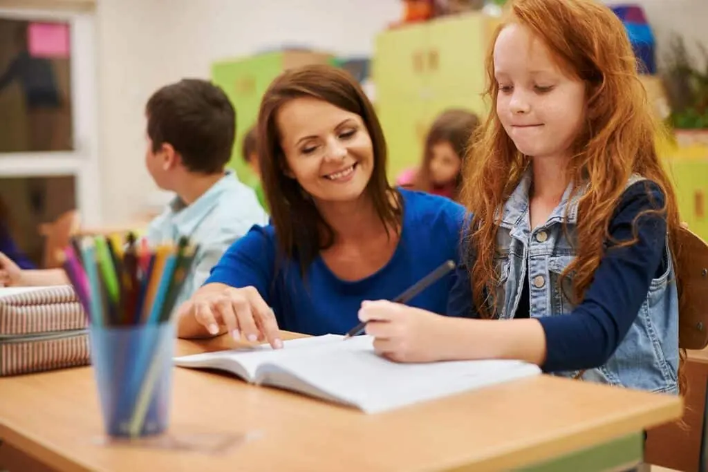 A teacher assisting a student sitting at a desk with her schoolwork