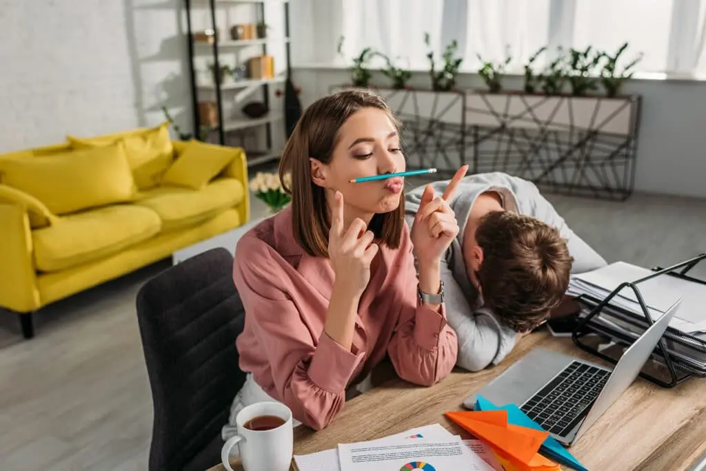 a girl holding a pencil between her nose and lip and a man sleeping on the table next to her.