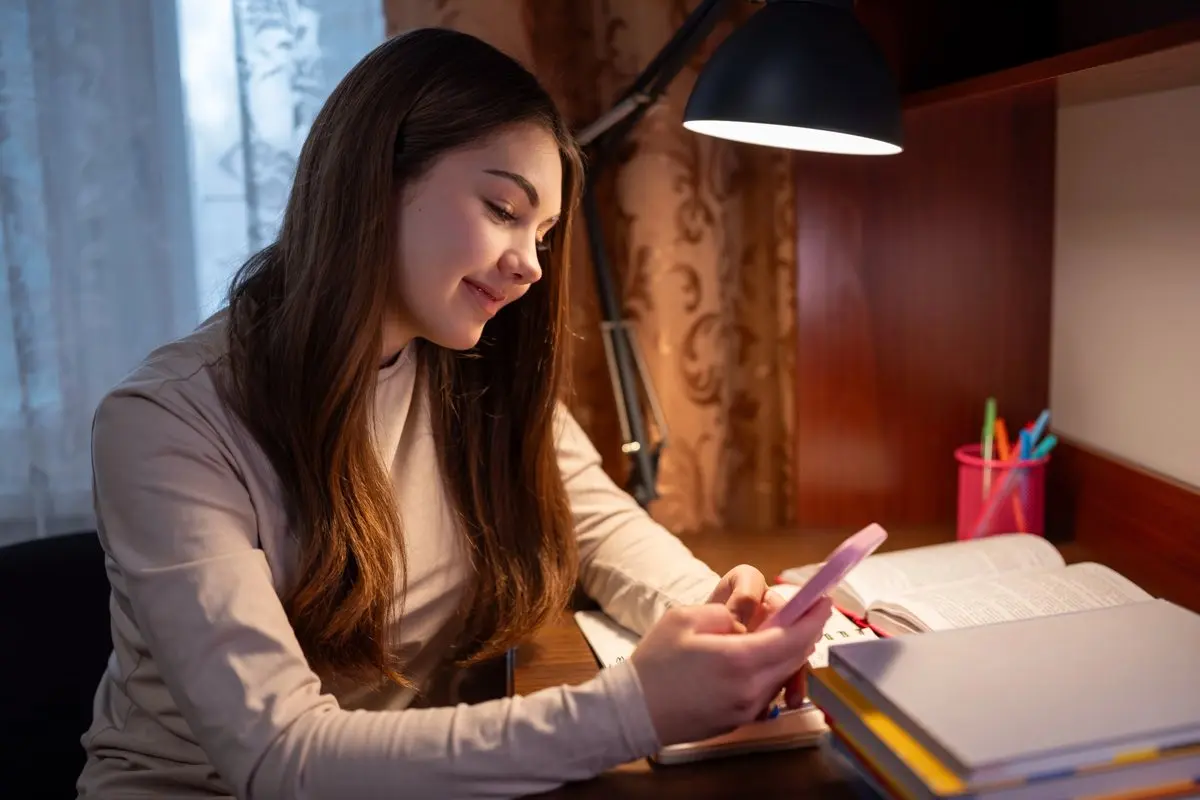 A student at her desk in the evening, looking at her phone instead of her open books